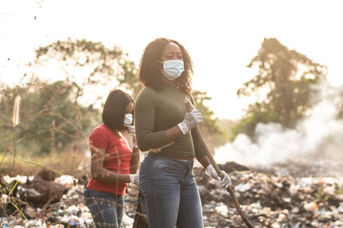 Two women with facemasks over their noses and mouths standing amidst a pile of smoking rubble/garbage